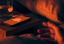 Celular sobre a mesa de cabeceira à noite com a tela acesa mostrando um sino de alerta, e uma mão indo pegá-lo