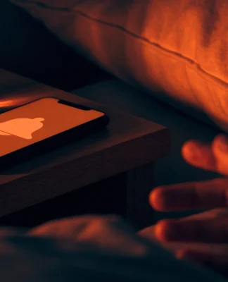 Celular sobre a mesa de cabeceira à noite com a tela acesa mostrando um sino de alerta, e uma mão indo pegá-lo