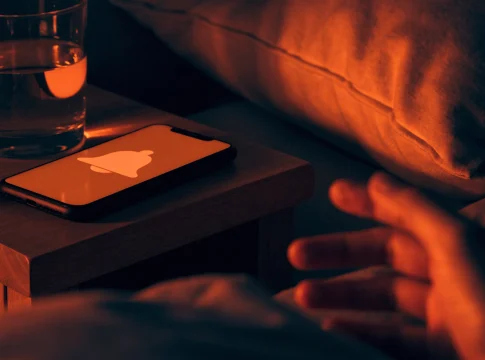 Celular sobre a mesa de cabeceira à noite com a tela acesa mostrando um sino de alerta, e uma mão indo pegá-lo