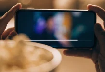 Celular na horizontal entre as duas mãos exibindo um vídeo, com uma tigela de pipoca desfocada à frente