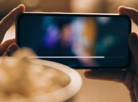 Celular na horizontal entre as duas mãos exibindo um vídeo, com uma tigela de pipoca desfocada à frente