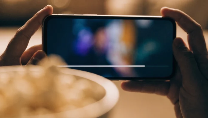 Celular na horizontal entre as duas mãos exibindo um vídeo, com uma tigela de pipoca desfocada à frente