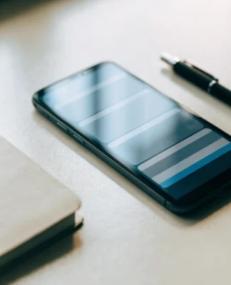 Celular deitado numa mesa clara ao lado de um caderno e uma caneta, exibindo uma lista