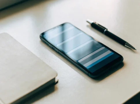 Celular deitado numa mesa clara ao lado de um caderno e uma caneta, exibindo uma lista