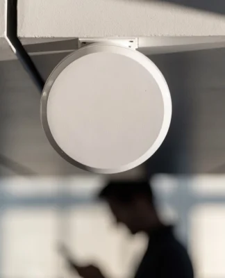 Ponto de acesso Wi-Fi branco e redondo fixado no teto, com uma pessoa desfocada usando o celular abaixo