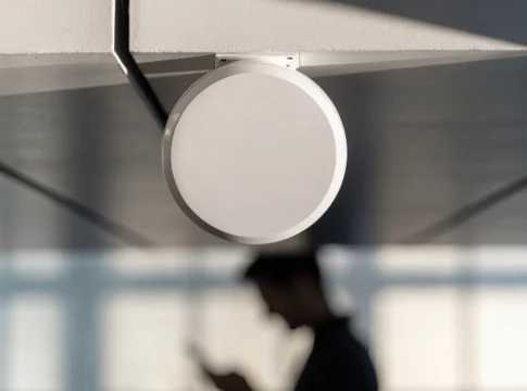 Ponto de acesso Wi-Fi branco e redondo fixado no teto, com uma pessoa desfocada usando o celular abaixo
