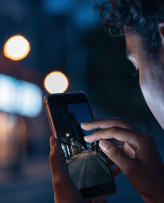 Pessoa fotografando uma rua escura à noite com o celular, e a tela mostra a cena bem mais clara