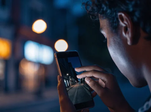 Pessoa fotografando uma rua escura à noite com o celular, e a tela mostra a cena bem mais clara