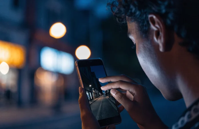 Pessoa fotografando uma rua escura à noite com o celular, e a tela mostra a cena bem mais clara