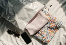 Embalagem branca aberta sobre a cama com roupas dobradas, óculos de sol e um celular