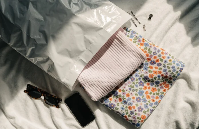 Embalagem branca aberta sobre a cama com roupas dobradas, óculos de sol e um celular